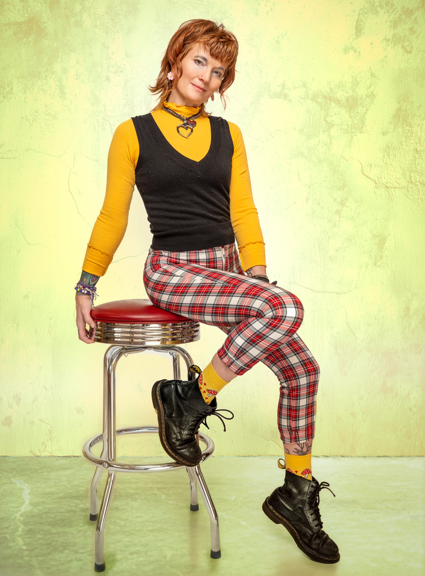 A woman wearing a long-sleeved yellow shirt, black vest, red plaid pants, and black boots, sits atop a red and chrome stool, posing against a cream-colored backdrop for a photograph
