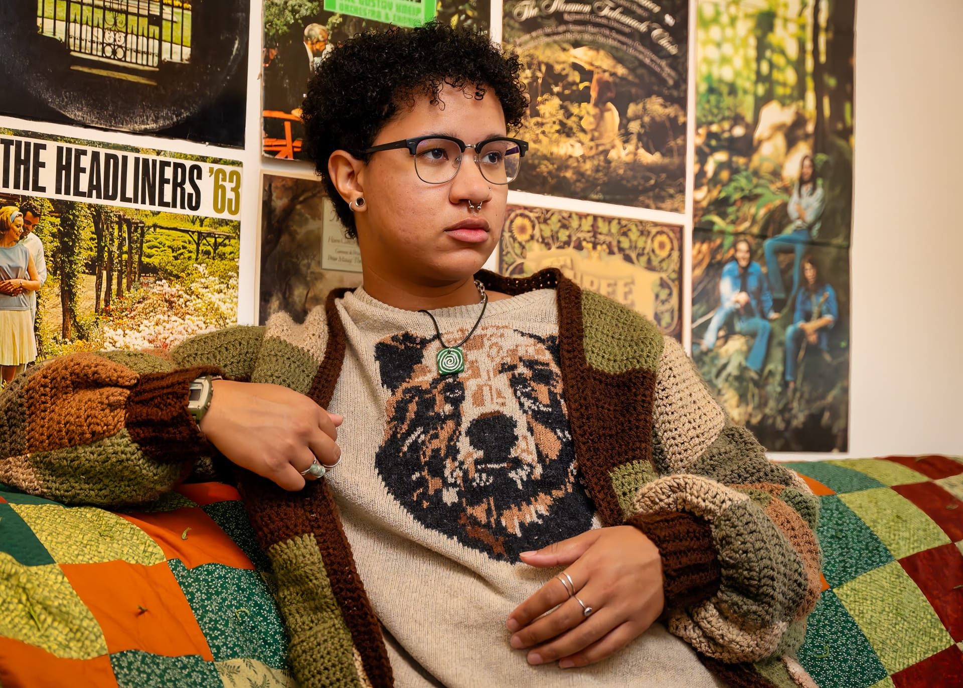 a mixed-race individual wearing an earth-tone cardigan over a knit sweater with bear motif sits on a quilt-draped couch. Vintage record album covers adorn the background wall