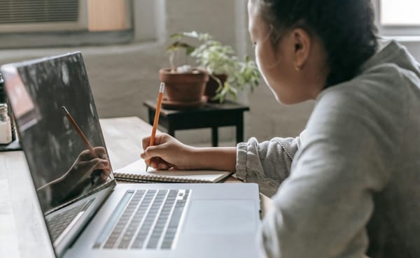 A teenage girl writes with a pencil in a notebook while sitting at a desk, a laptop open in front of her.