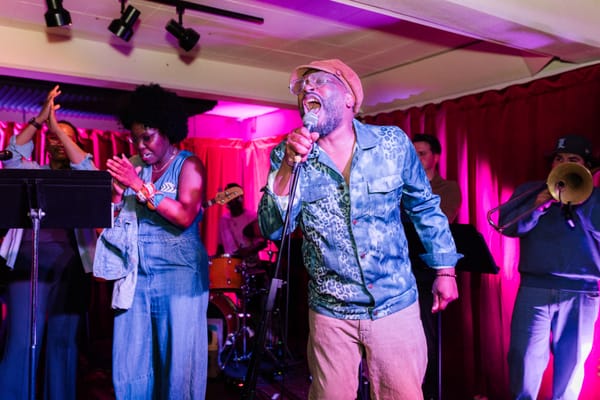 A Black man sings loudly into a microphone as two Black women clap along; a band plays in the background