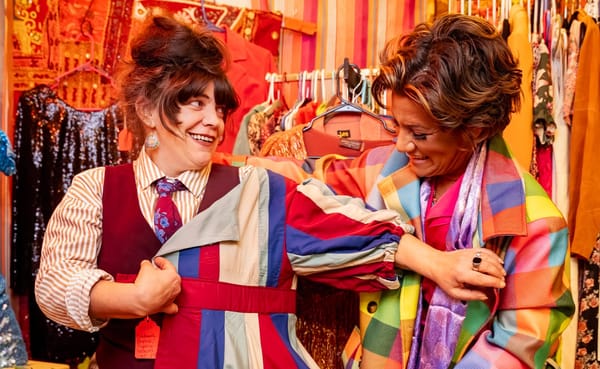 two women try on brightly colored jackets against a red and orange backdrop